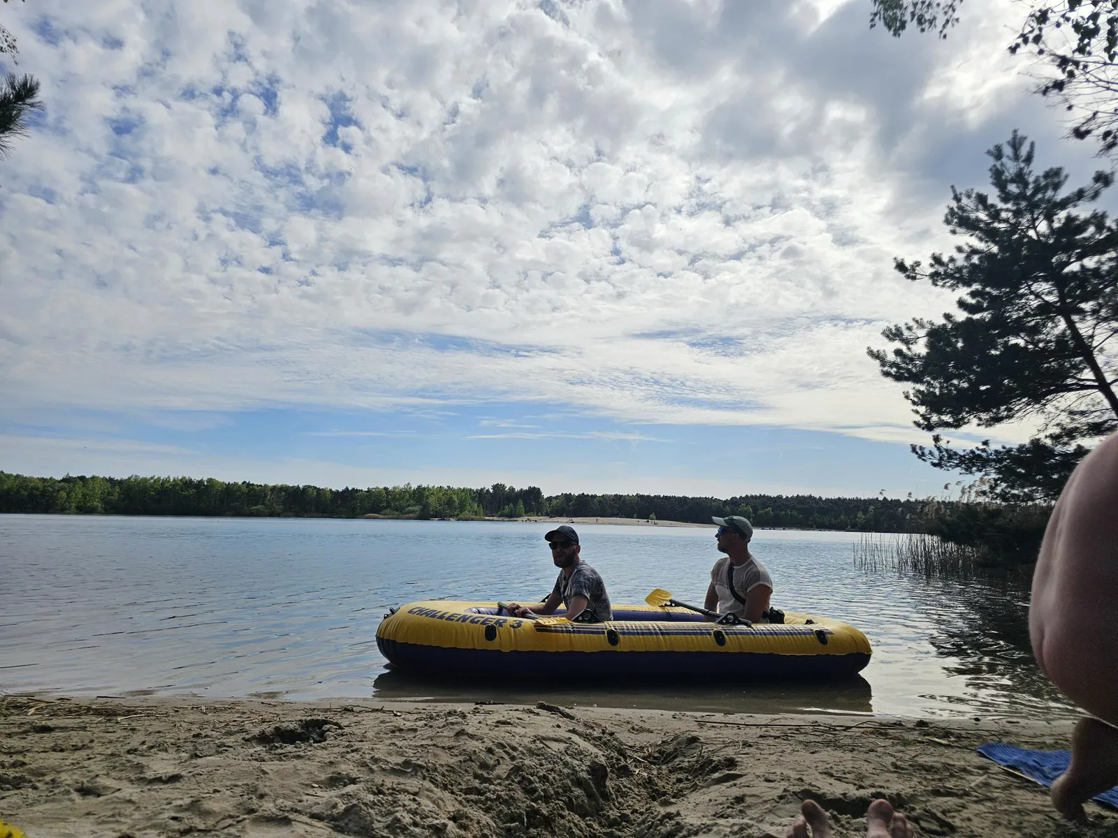 A wide lake under a bright cloudy sky, with a small inflatable boat drawn up on the sandy shore.