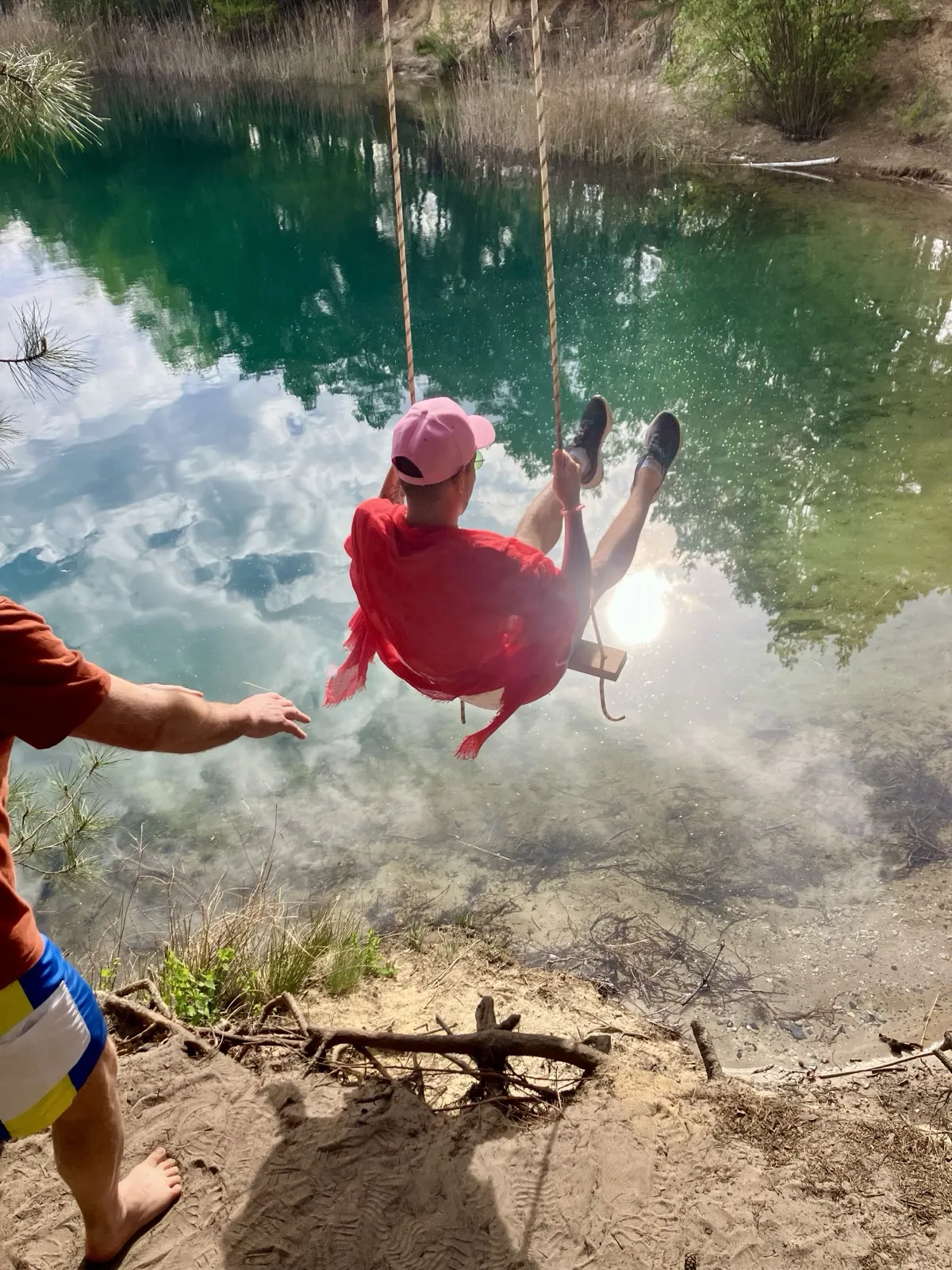 A faerie in a red robe swinging out over a green lake on a rope swing, being given a push from the bank.