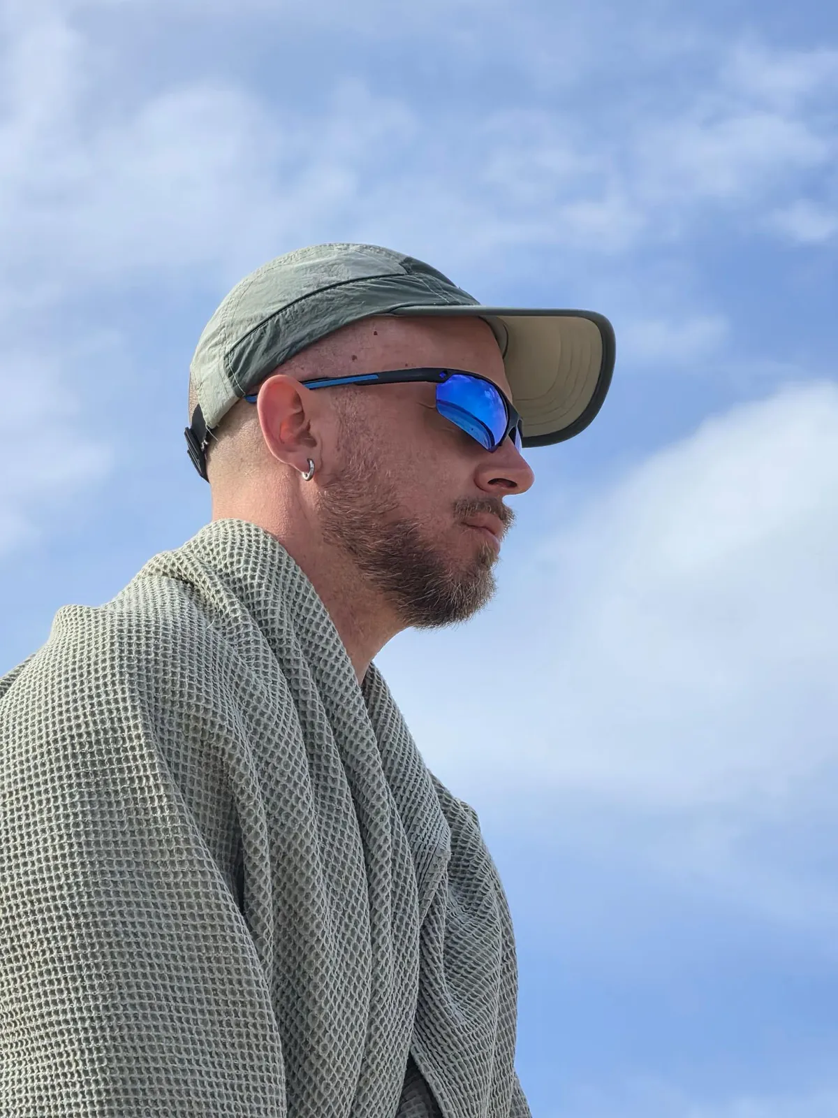 A faerie in a cap and sunglasses with a shawl around their shoulders, looking out across a wide blue sky.